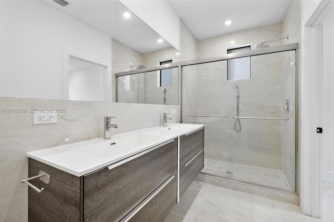 Floating double vanity with a white countertop and dual chrome faucets, complemented by a large wall-to-wall mirror, walk-in shower with sliding glass doors, and light-toned tile flooring