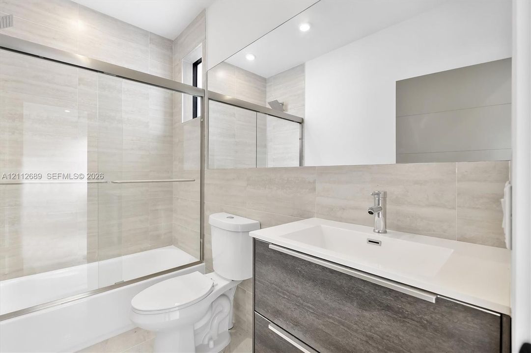 Bathroom featuring a wall-to-wall mirror, modern floating vanity with a stone-top integrated sink, floor-to-ceiling tile surround, and a glass-enclosed tub shower with metallic hardware