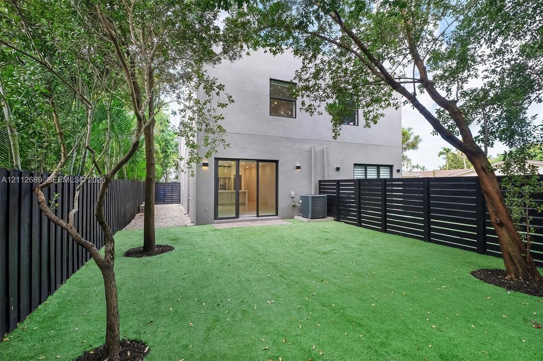Fenced backyard featuring synthetic turf, mature trees, smooth stucco exterior, and sliding glass doors