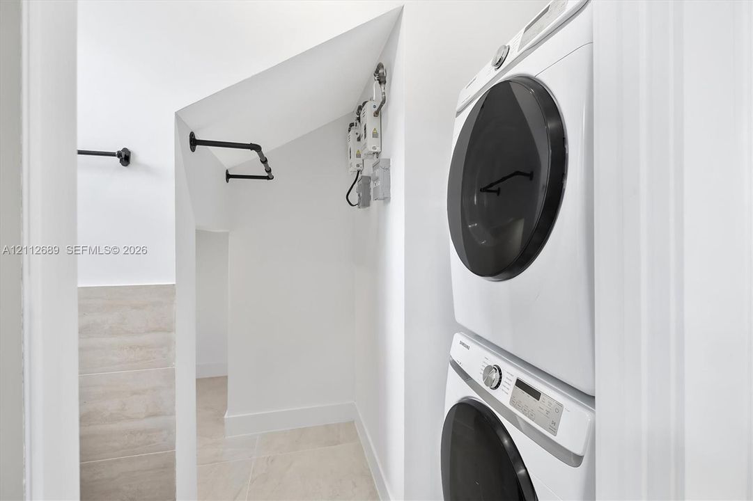 Laundry area featuring stacked appliances, matte black wall-mounted metal shelving, neutral large-format floor tiling, and white wall finishes