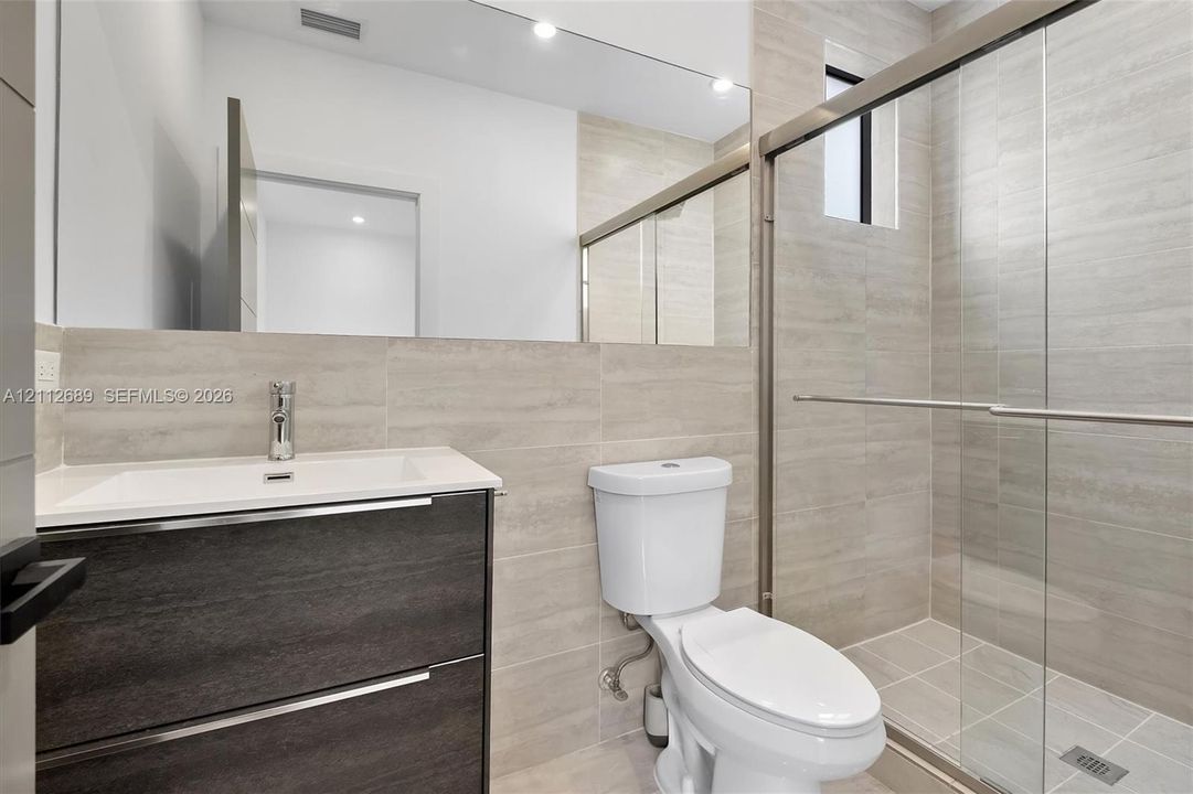 Wall-mounted vanity with a rectangular sink, full-width mirror, walk-in shower with sliding glass doors, and neutral large-format tile wall cladding