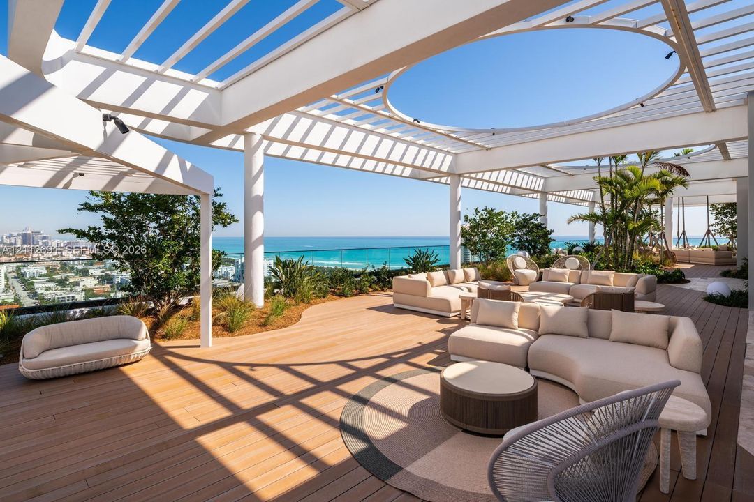 Expansive rooftop terrace featuring a white pergola structure with a circular cutout, wood-finish decking, glass panel railings, and perimeter landscaping