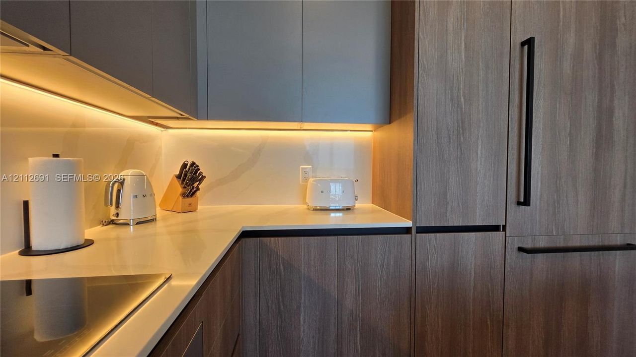 Kitchen featuring wood-finish cabinetry, solid surface countertops, a full-height backsplash, under-cabinet lighting, and integrated panel-ready appliances