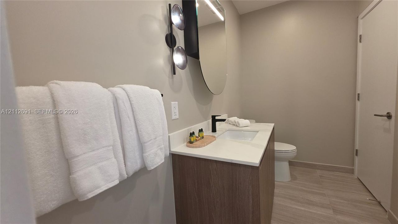 Modern vanity with wood-finish cabinetry, white countertop, integrated rectangular sink, matte black faucet, and oval mirror with wall-mounted lighting