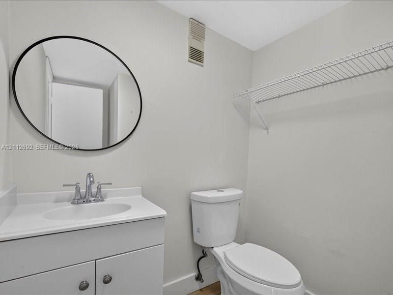 White vanity with an integrated countertop, oval mirror featuring a matte black frame, dual-handle faucet, and white wall-mounted wire shelving