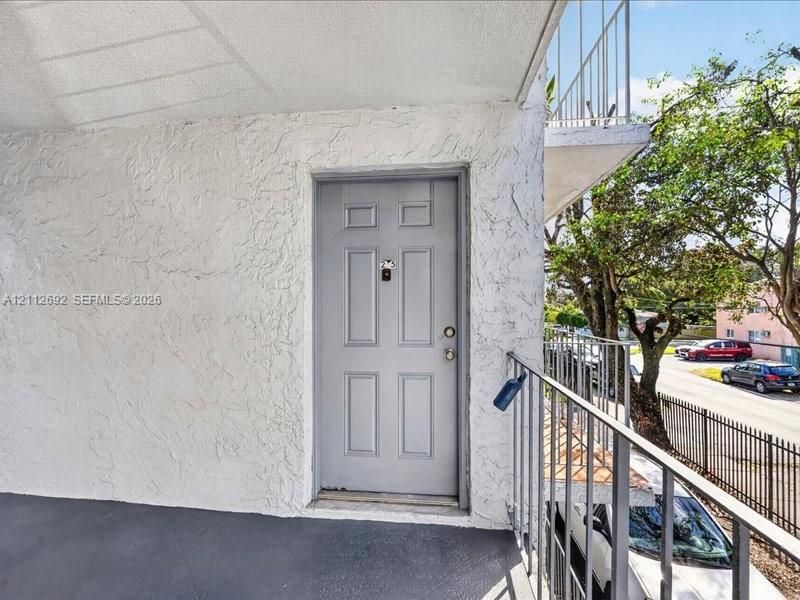 Exterior entry featuring a textured stucco facade, a paneled door with hardware, and a metal railed walkway