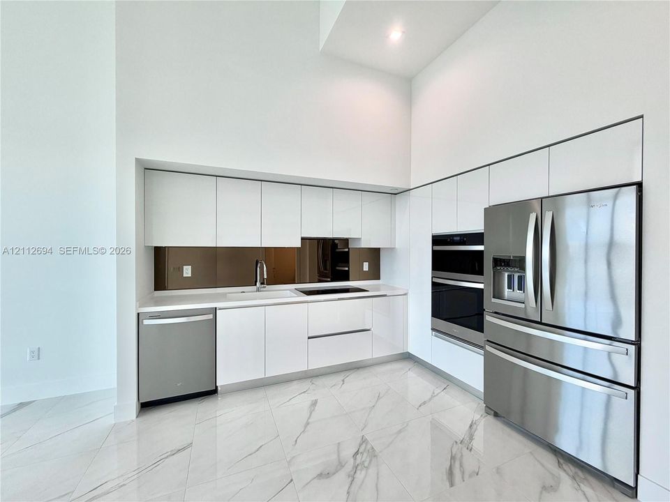 Kitchen featuring high-gloss white cabinetry, stainless steel appliances, built-in dual wall ovens, solid-surface countertops, and large-format porcelain tile flooring