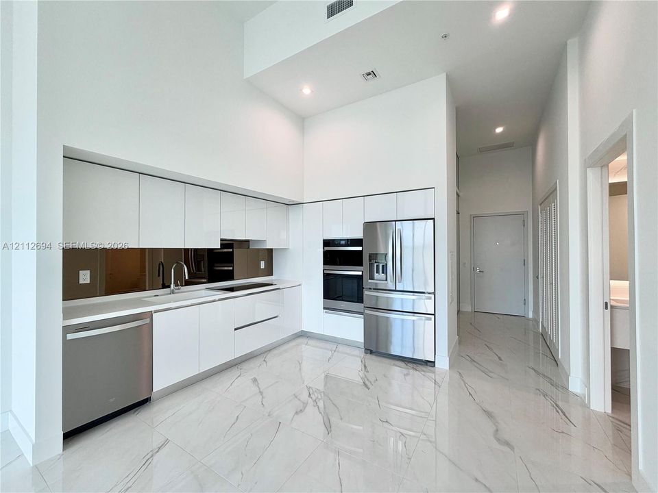 High-gloss white cabinetry, polished stone-finish flooring with veining, stainless steel built-in appliances, a mirrored backsplash, and high ceilings