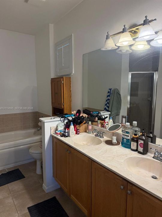 Dual-sink vanity with a solid-surface countertop and light-wood cabinetry, large wall-mounted mirror, four-light vanity fixture, built-in medicine cabinet, and a tiled soaking tub