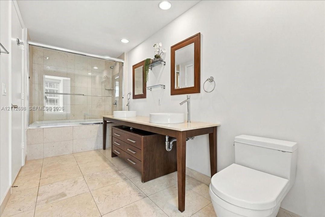 Double vanity with dual vessel sinks, stone-surface countertop, dark wood-finish cabinetry, and a glass-enclosed shower with tile surround