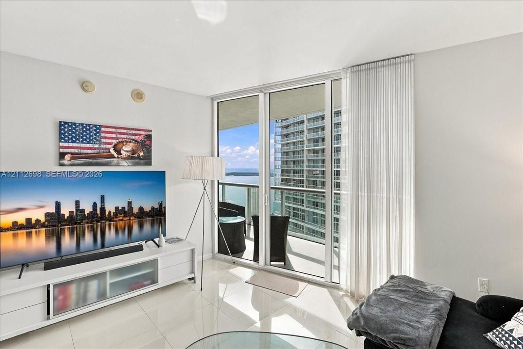 Floor-to-ceiling glass sliding doors, polished tile flooring, and a covered balcony overlooking the water and metropolitan skyline