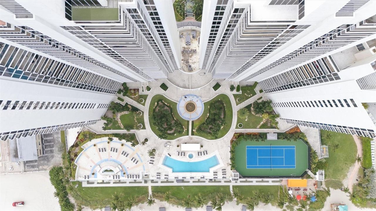 Aerial perspective of high-rise condominium complex featuring a central courtyard with structured landscaping, a swimming pool, a tennis court, and paved pedestrian paths