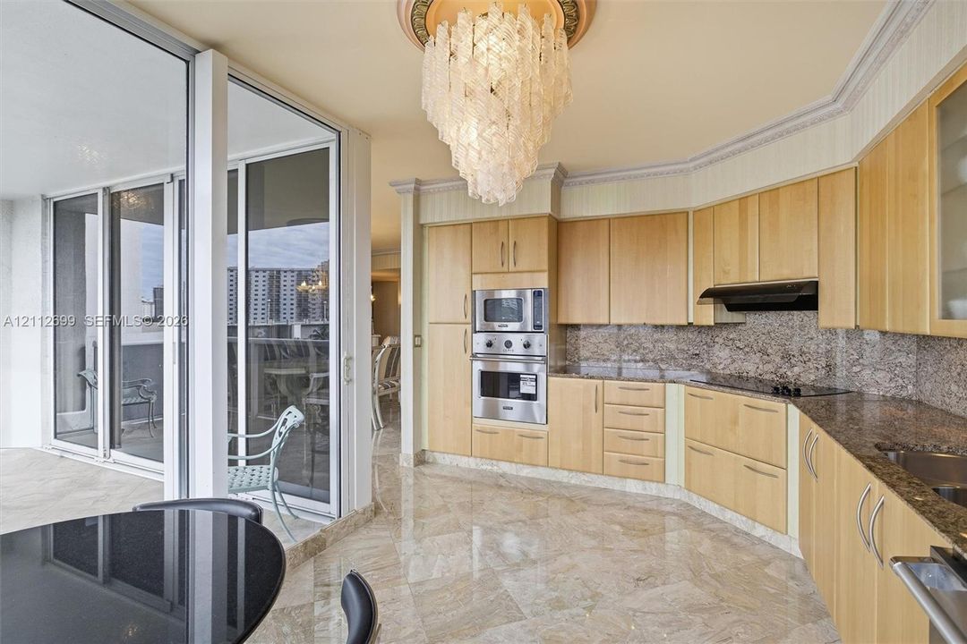 Kitchen featuring light-finish cabinetry, stone countertops with a matching full-height backsplash, polished stone flooring, integrated stainless steel appliances, and a decorative crystal chandelier with an ornate ceiling medallion