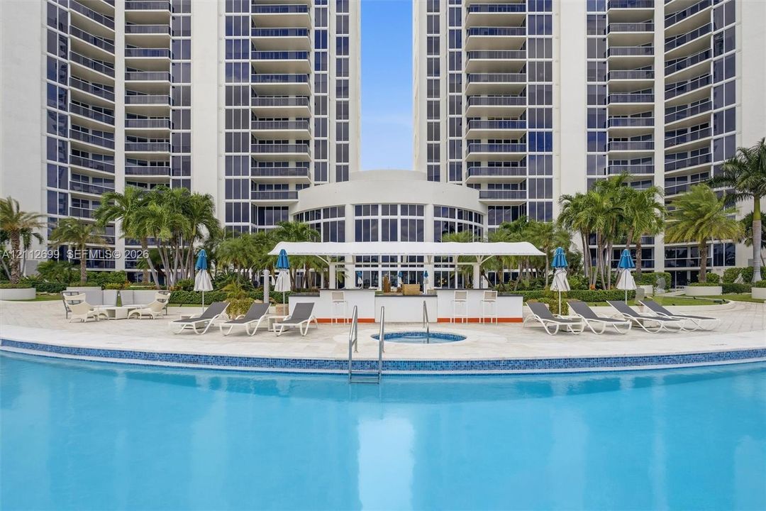 Curved swimming pool with tile trim, integrated spa, poolside bar under a covered canopy, and paved deck featuring mature palm trees