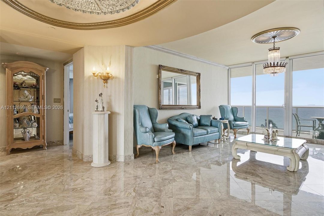 Polished stone flooring, recessed tray ceiling with gold-tone molding, crystal chandelier, and floor-to-ceiling glass sliding doors with ocean backdrop