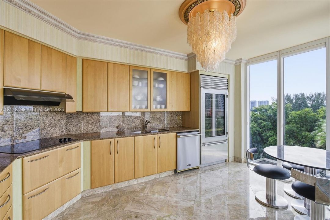 Kitchen featuring light wood cabinetry, stone countertops, stone flooring, built-in stainless steel appliances, and a tiered glass chandelier
