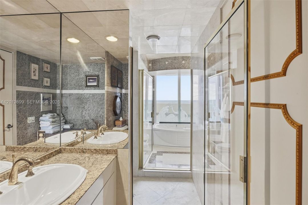 Bathroom featuring a stone-topped dual vanity with integrated sinks, floor-to-ceiling mirrors, tile wall detailing, a jetted soaking tub, and custom paneled doors with gold-tone trim