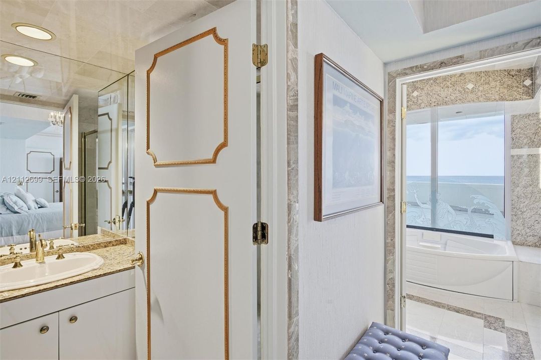 Vanity featuring an oval undermount sink, stone countertop, and full-height mirrored wall, paired with a jetted soaking tub positioned against a large window with ocean scenery