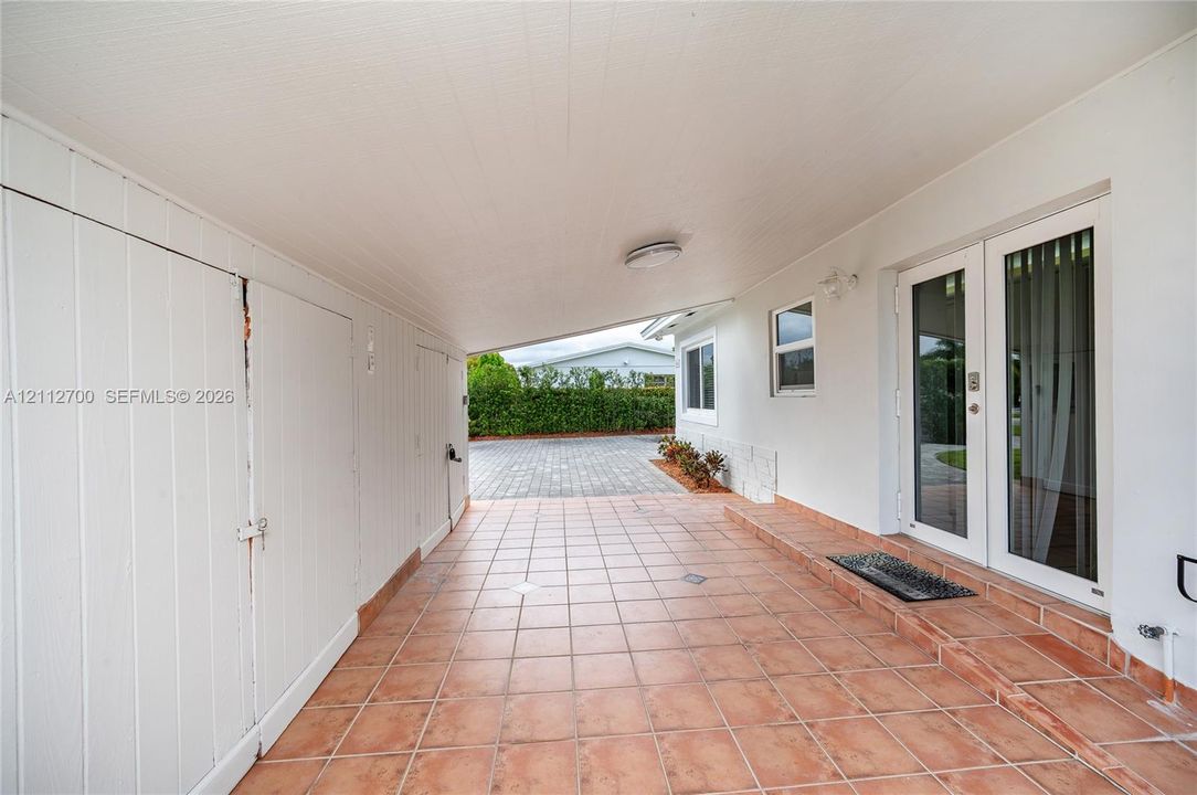 Covered carport featuring terracotta-style tile flooring, built-in storage cabinetry, a double-door entrance with glass inserts, and stone-paver driveway access
