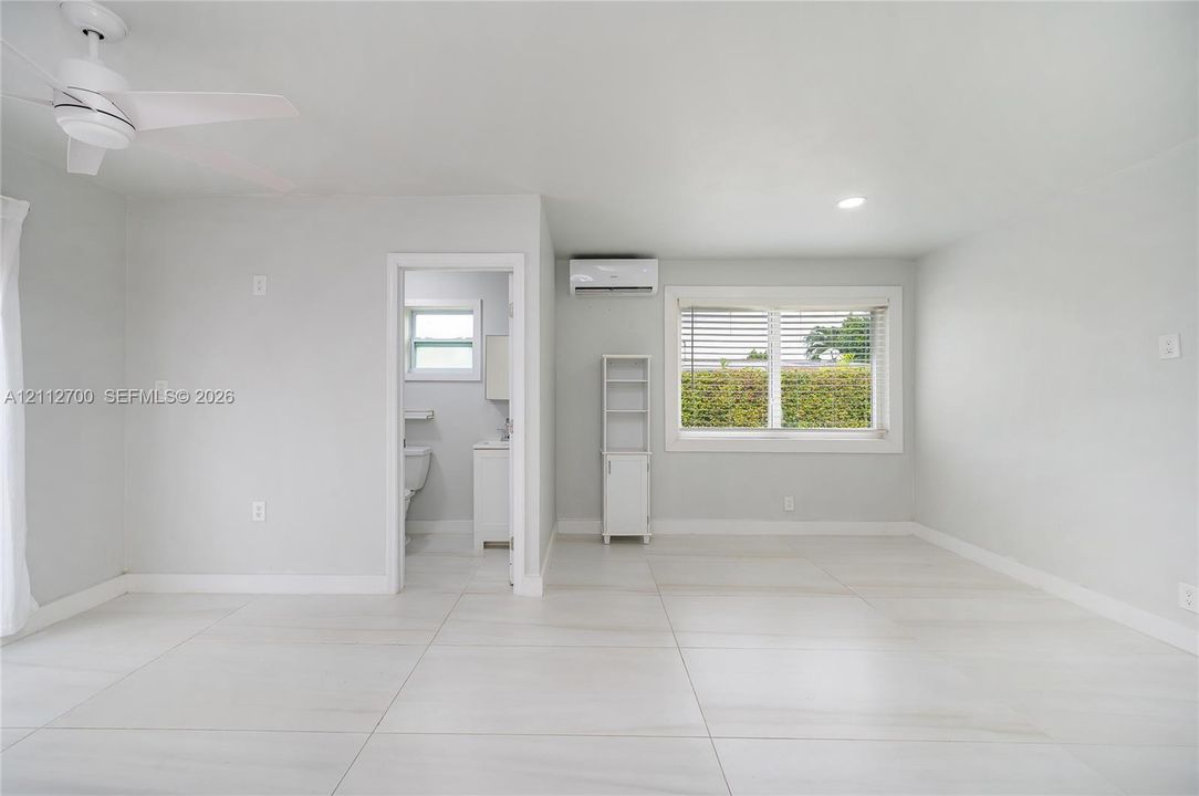 Large-format tile flooring, ceiling fan with integrated lighting, wall-mounted ductless air conditioning unit, and expansive window with horizontal blinds