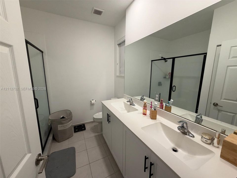 Dual-sink vanity with white cabinetry, quartz-style countertops, matte black hardware, and an expansive wall mirror, complemented by a walk-in shower with a framed glass enclosure