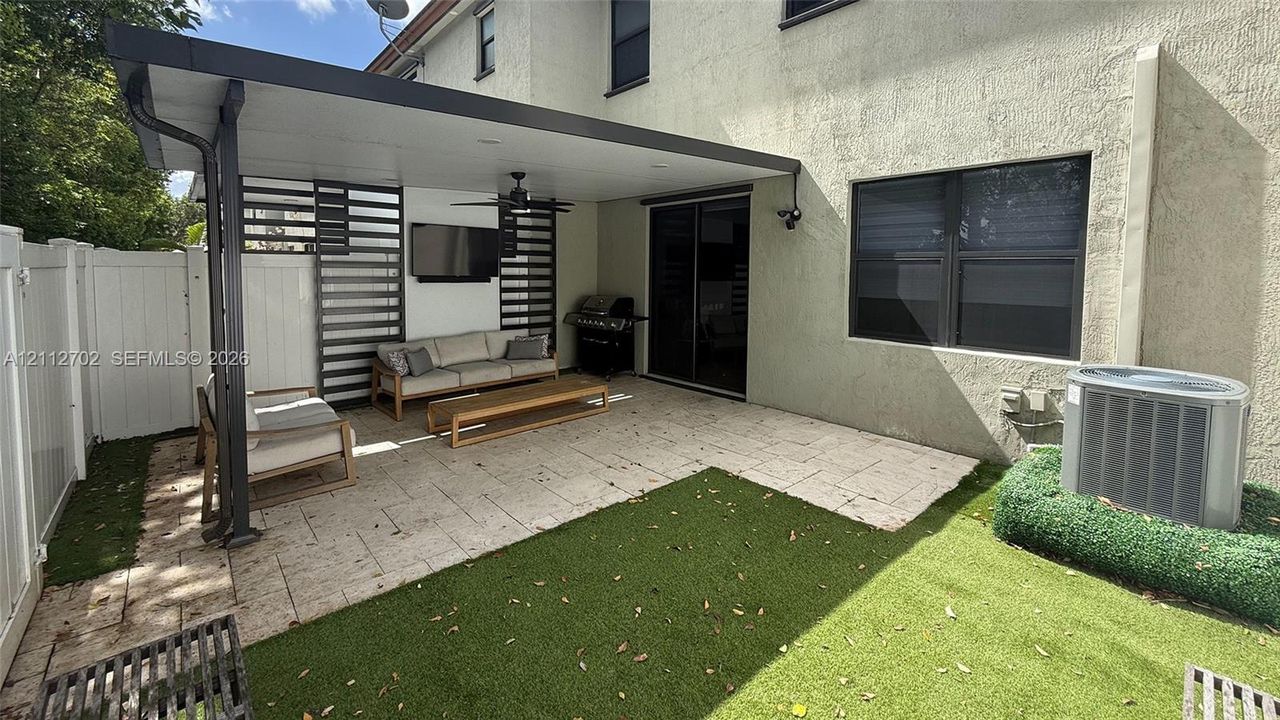 Covered patio featuring a solid roof structure with a ceiling fan, stone-style pavers, horizontal privacy screening, and synthetic turf lawn area