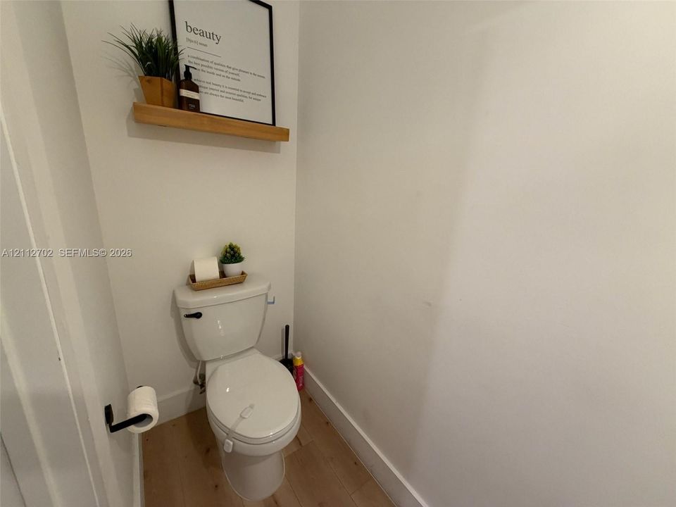 Standard toilet featuring a matte black flush handle and wall-mounted matte black toilet paper holder, complemented by a light wood-finish floating shelf and wood-finish flooring