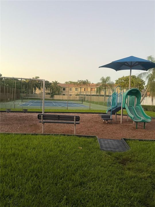 Recreational grounds featuring a fenced tennis court, a playground structure with slides, a swing set, and a metal bench