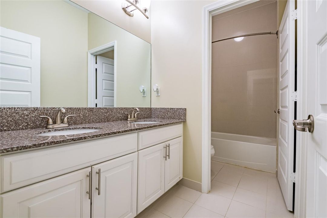 Bathroom featuring a dual-sink vanity with stone-composite countertop, white cabinetry with brushed-metal pulls, full-width vanity mirror, and a tub-shower combination with tiled surround