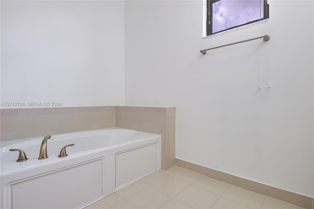 Built-in soaking tub with a panelled apron front, neutral tile surround, metallic deck-mounted faucet, tile flooring, and a wall-mounted towel bar