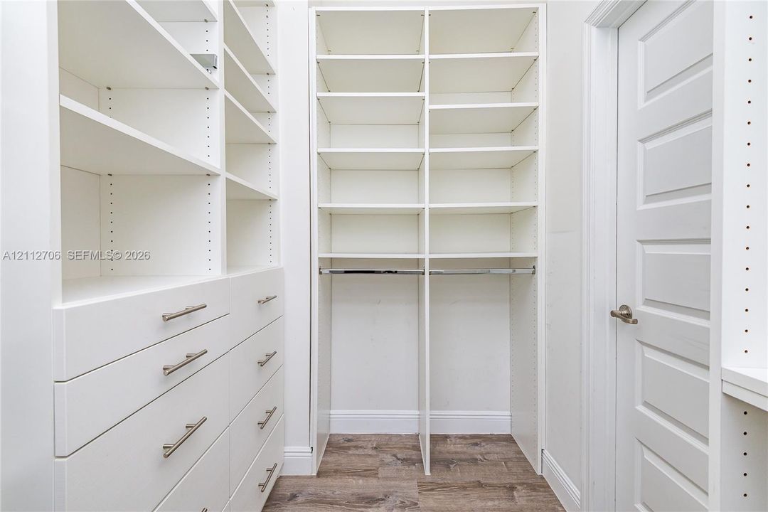 Custom walk-in closet featuring white built-in shelving, floor-to-ceiling storage units, multiple drawers with satin-finish pulls, integrated hanging rods, and wood-finish flooring