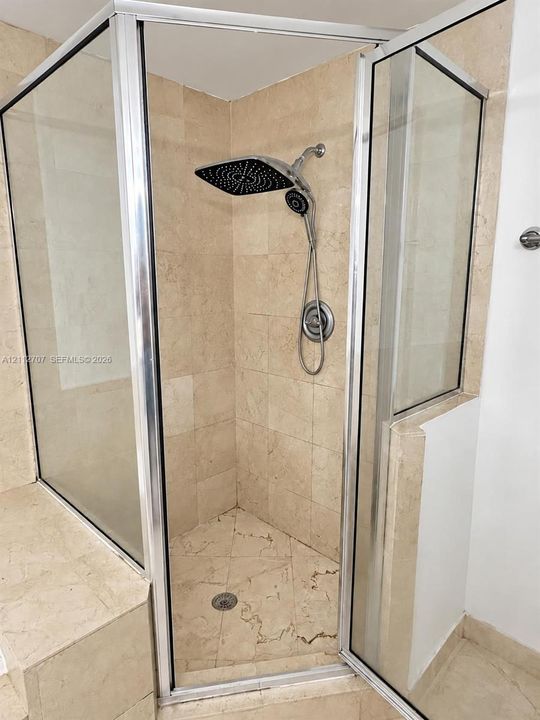Corner shower enclosure featuring floor-to-ceiling stone-tiled walls and flooring, a built-in stone bench, a rainfall shower head with a handheld attachment, and a chrome-framed glass door