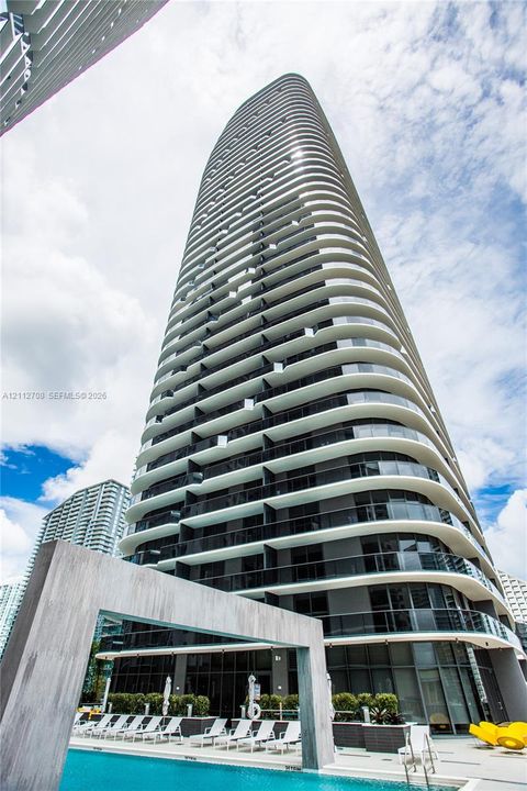 High-rise residential tower featuring curved exterior balconies, floor-to-ceiling glass windows, and a ground-level swimming pool with waerfall
