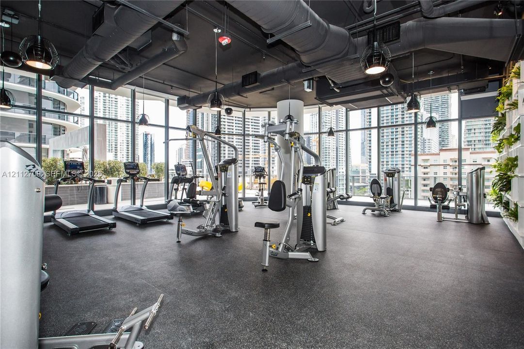 Fitness center featuring floor-to-ceiling windows, exposed industrial-style ductwork, recessed pendant lighting, and a vertical plant wall