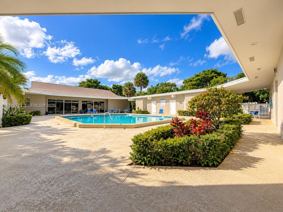 In-ground swimming pool surrounded by a spacious textured concrete patio, complemented by a manicured shrub landscape and a covered exterior walkway