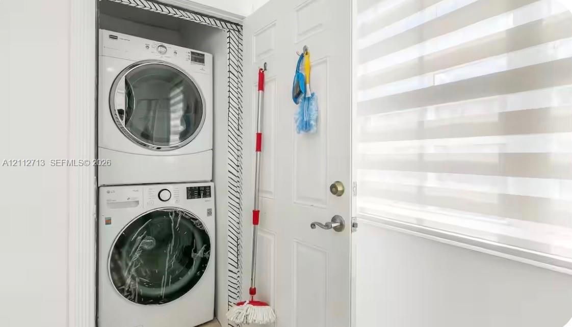 Stacked laundry unit featuring a front-loading washer and dryer, housed within a closet featuring patterned interior trim and a multi-panel door with metallic hardware, accompanied by horizontally-striped roller blinds