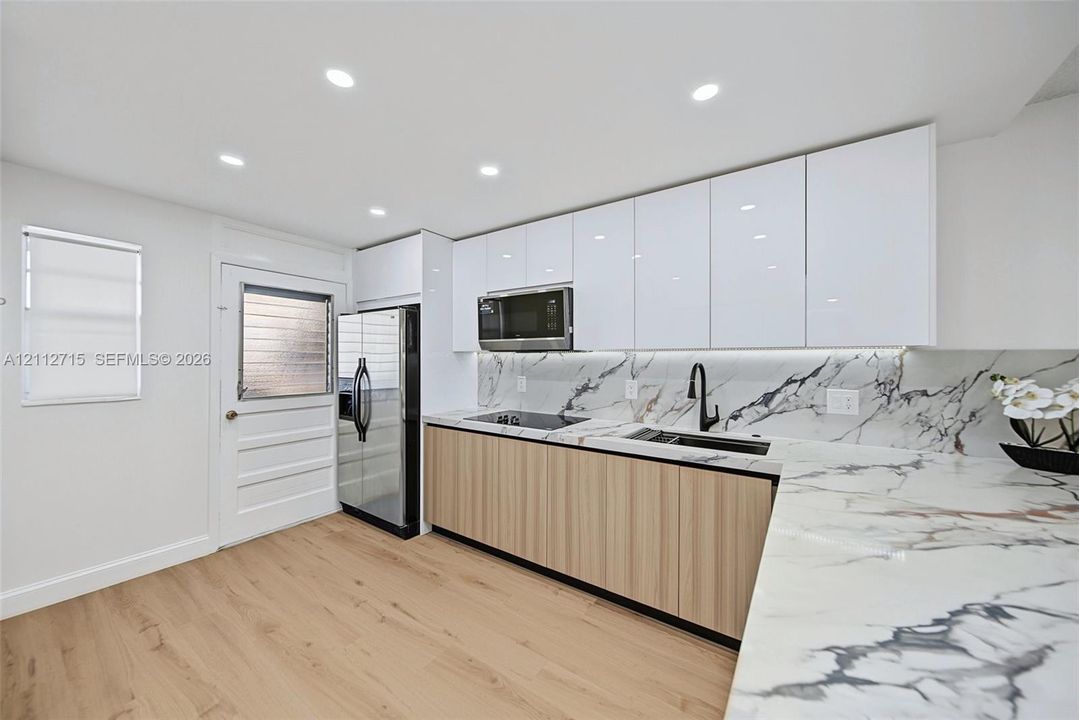 Contemporary kitchen featuring high-gloss white upper cabinetry, wood-finish lower cabinetry, stone-patterned countertops and backsplash, integrated stainless steel appliances, and a matte black gooseneck faucet