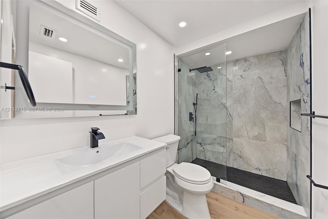 Bathroom featuring a floating vanity with a white countertop, wall-mounted illuminated mirror, walk-in shower with a glass enclosure, floor-to-ceiling stone-patterned wall tile, and matte black fixtures