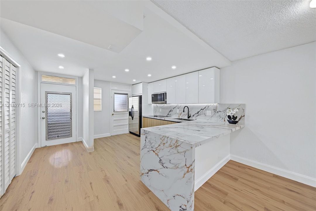 Kitchen featuring white high-gloss upper cabinetry, wood-finish base cabinetry, waterfall-edge countertop, stone-finish backsplash, and recessed lighting