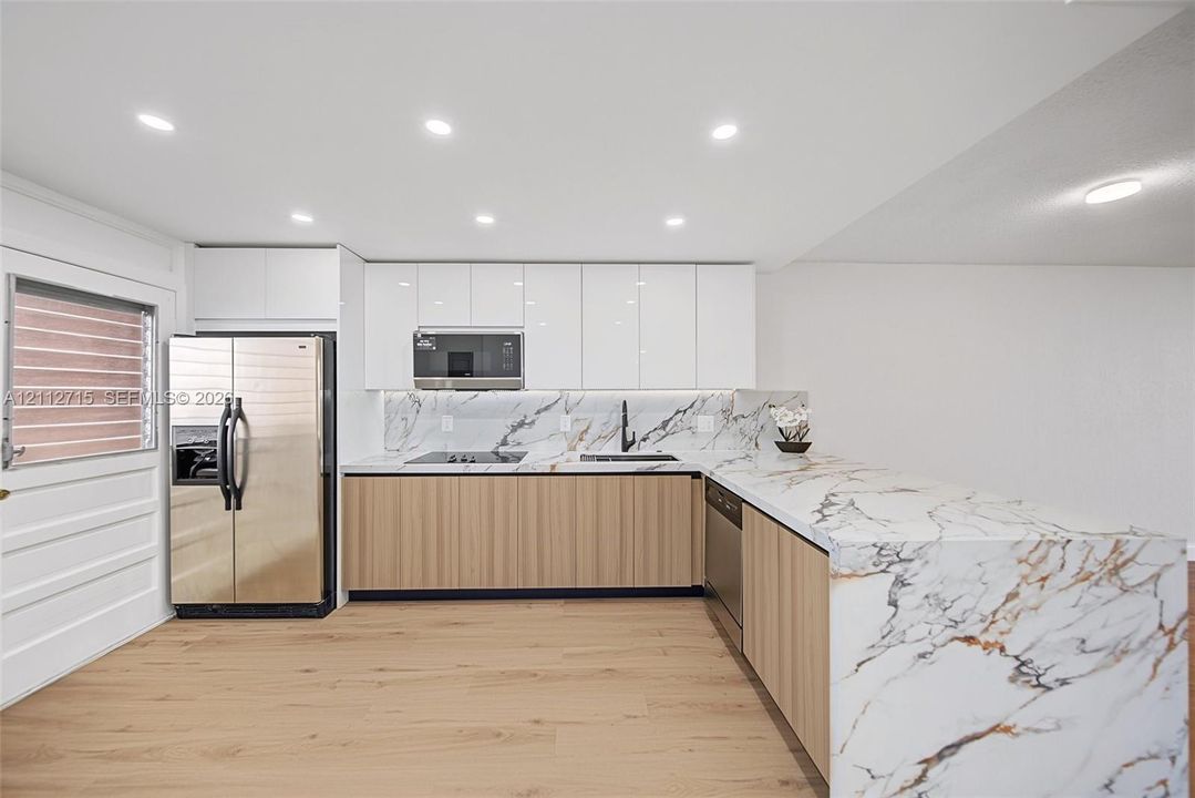 Kitchen featuring high-gloss white upper cabinetry, wood-finish lower cabinetry, stone-patterned countertops with matching backsplash, stainless steel appliances, and recessed lighting
