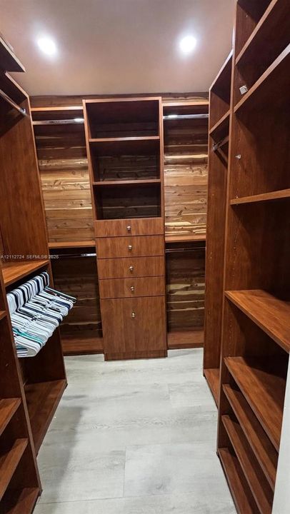 Walk-in closet featuring floor-to-ceiling wood-finish custom cabinetry, a central drawer stack, integrated hanging rods, open shelving, and light-tone wood-finish flooring