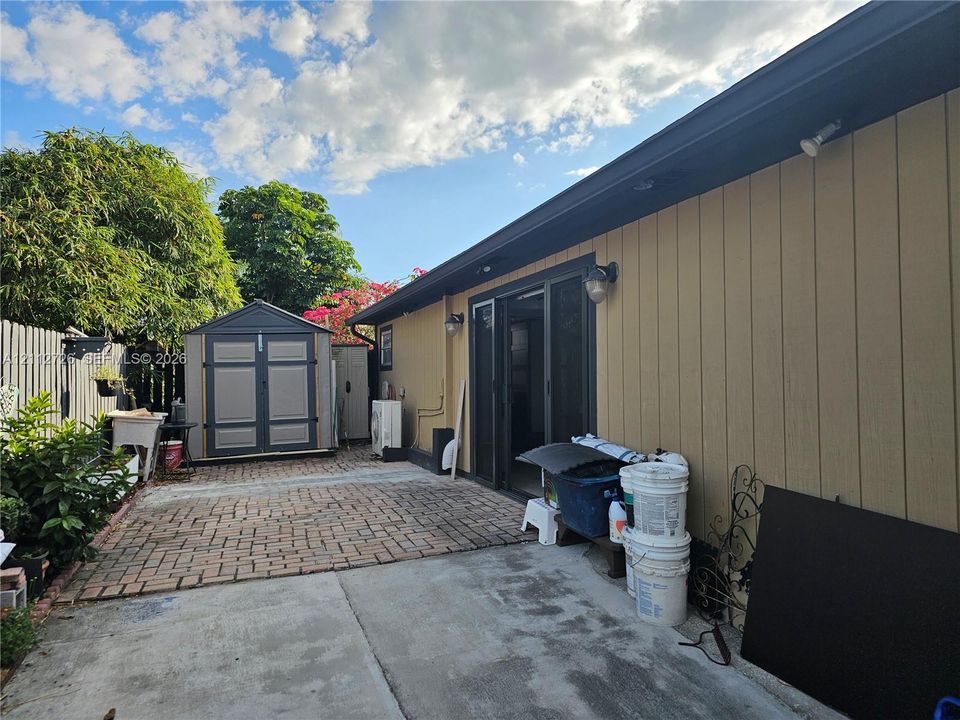 Exterior space featuring vertical siding, brick-patterned hardscaping, sliding glass doors, attached exterior lighting, and a detached storage structure