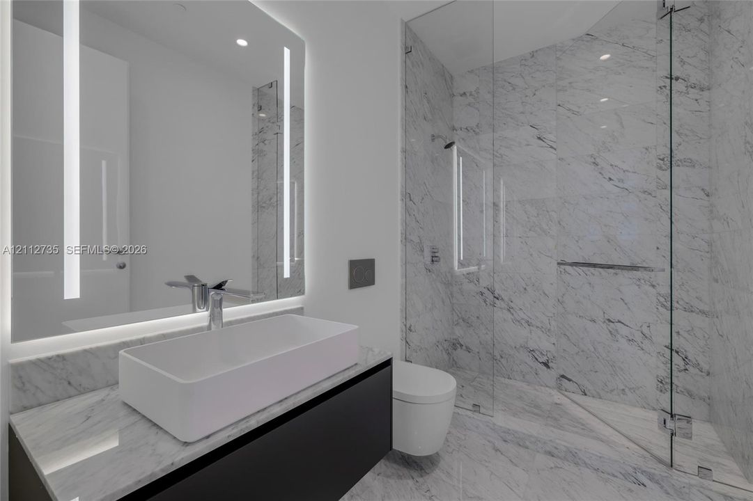 Floating vanity with integrated countertop, rectangular vessel sink, wall-to-wall mirror with vertical integrated lighting, glass-enclosed shower, and stone-finish wall and floor tiling