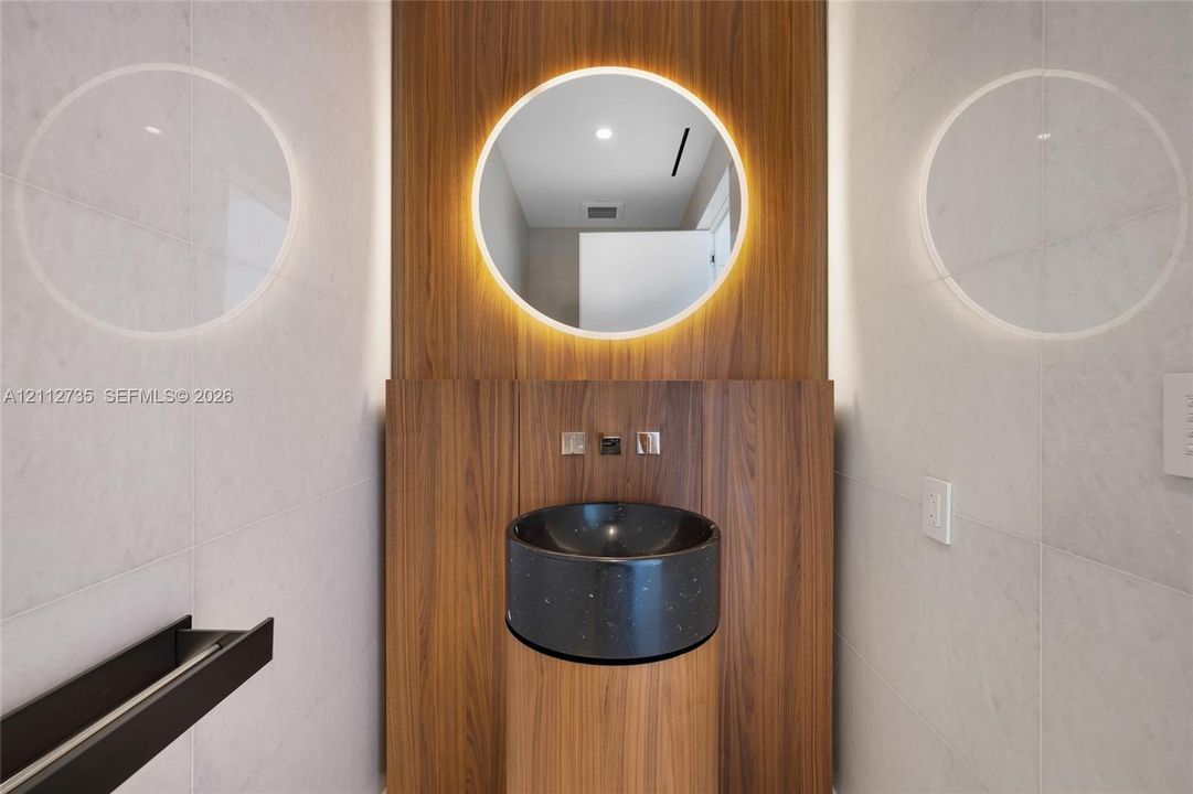Wall-mounted wood-finish paneling, circular backlit mirror, cylindrical wood-finish pedestal vanity, black stone vessel sink, and wall-mounted metallic fixtures
