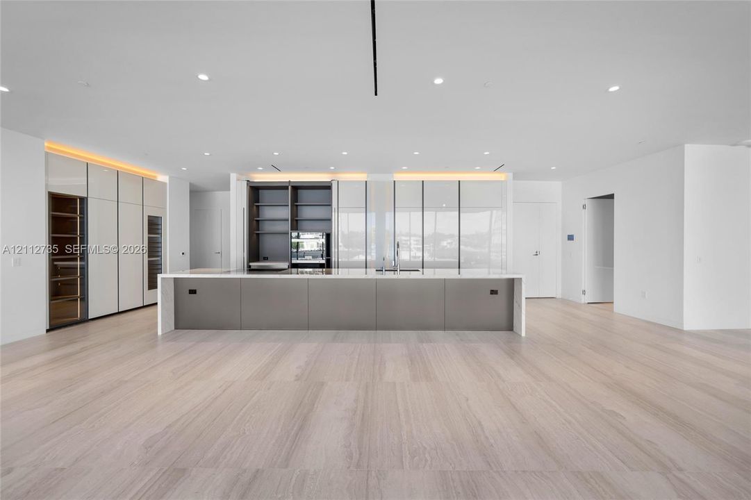 Minimalist kitchen featuring an expansive center island with a stone countertop, floor-to-ceiling cabinetry with integrated display shelving, built-in wine refrigeration, and light-toned wood-finish flooring
