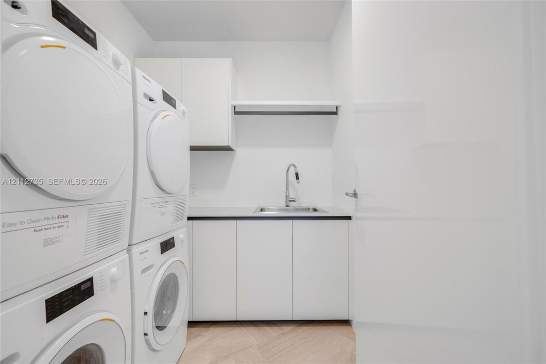 Utility area featuring stacked laundry appliances, white built-in cabinetry, a rectangular stainless steel sink with a high-arch pull-down faucet, and light-tone wood-finish flooring