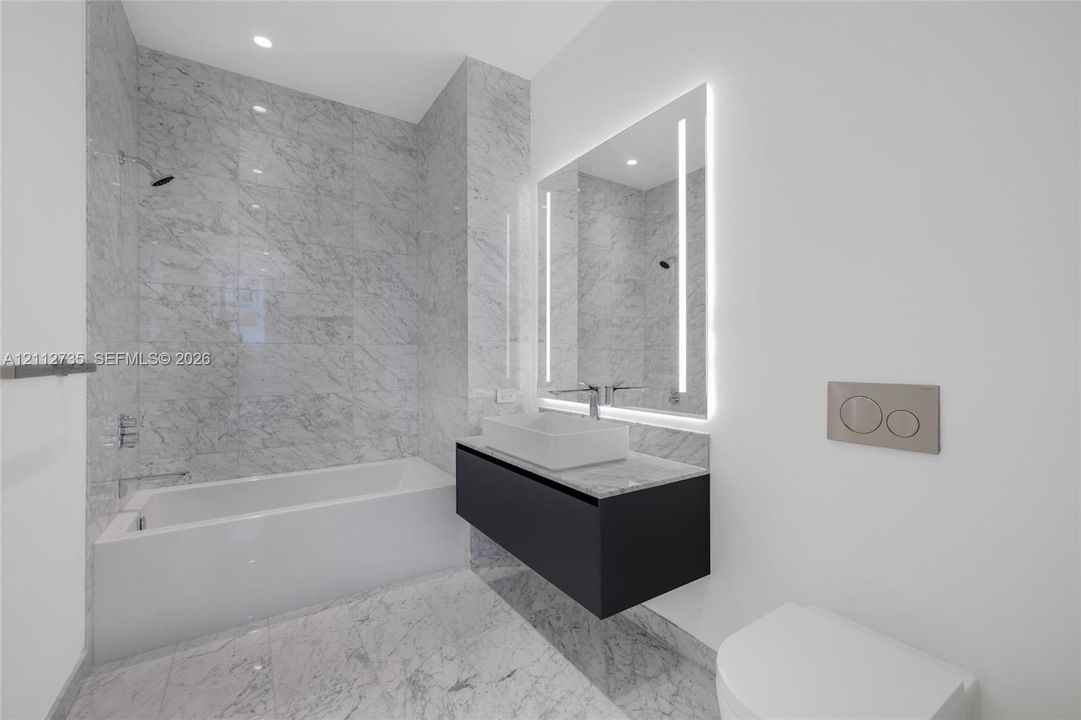 Full-height stone wall tiling, floating vanity with vessel sink, wall-mounted faucet, illuminated mirror, and wall-mounted dual-flush toilet