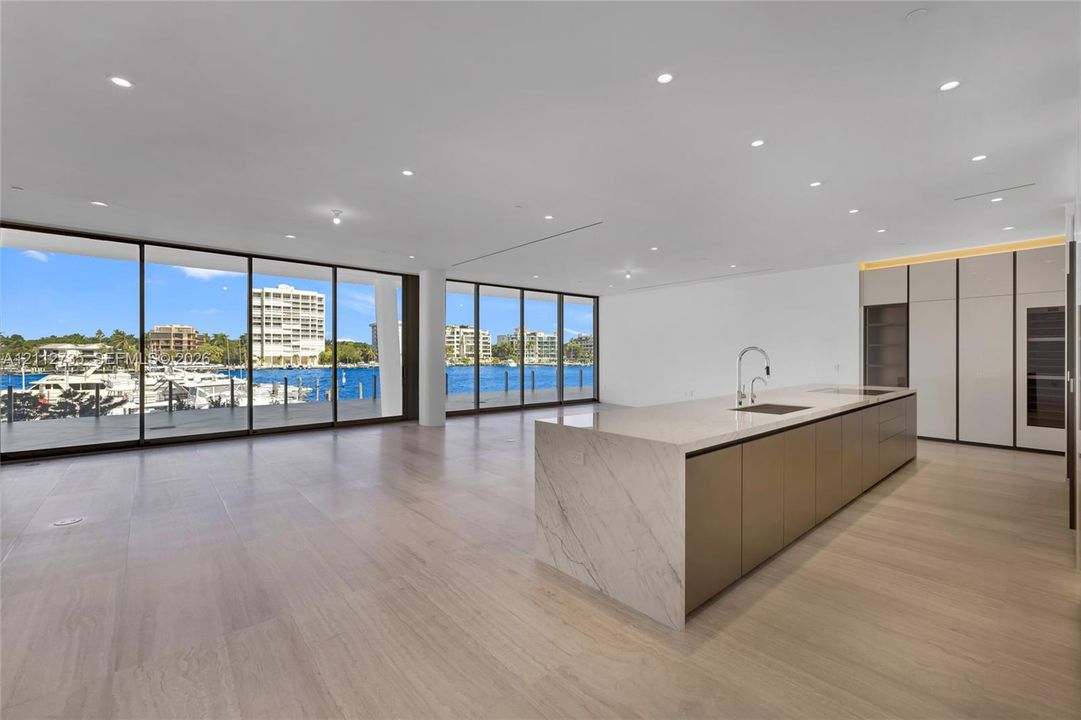 Open-concept interior featuring floor-to-ceiling glass walls, expansive kitchen island with a stone-finish waterfall countertop, built-in floor-to-ceiling cabinetry, and light-toned wood-finish flooring