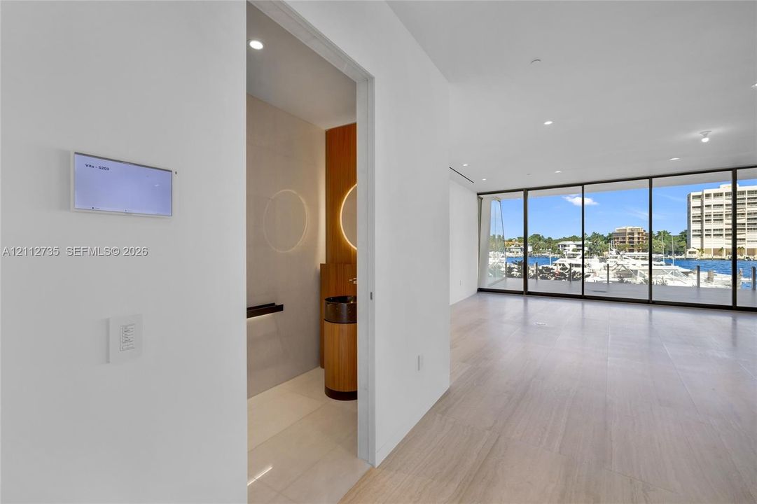 Floor-to-ceiling glass wall, wood-finish flooring, recessed lighting, and wall-mounted smart home control panel, featuring a powder room with cylindrical vanity and circular back-lit mirror
