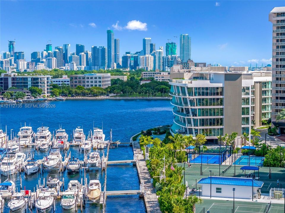 Waterfront property featuring a private marina, professional-grade tennis courts, and multi-level architectural glass facade with a metropolitan skyline backdrop